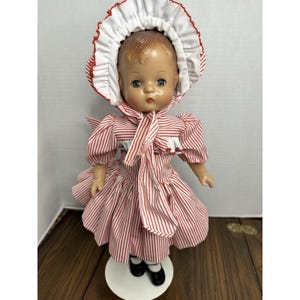 May include: A vintage doll with a painted face, wearing a red and white striped dress and matching bonnet. The doll has black shoes and is standing on a white stand. The dress has a bow at the neck.