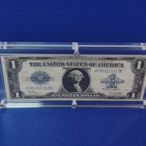 Peut inclure: Un billet d'un dollar présenté dans un boîtier en acrylique transparent. Le billet présente un portrait de George Washington et le texte "ONE SILVER DOLLAR". Le boîtier rectangulaire a des fixations argentées, sur fond bleu.
