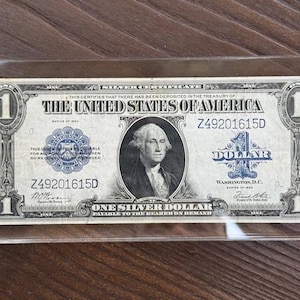 Puede incluir: Un billete de un dólar de plata de los Estados Unidos, con un retrato de George Washington. El billete está encerrado en un soporte protector transparente. El texto "ONE SILVER DOLLAR" es visible.