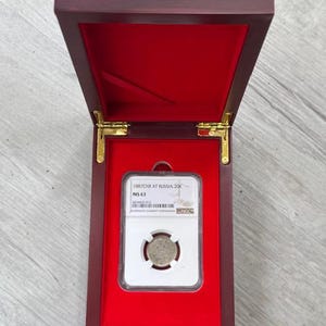 May include: A wooden display box with a red velvet interior holds a coin encased in a clear plastic holder. The box is open, revealing the coin and its protective case. The box has gold-coloured hinges.