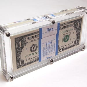 May include: A clear acrylic banknote holder with a stack of $1 bills inside. The holder has a blue label that reads "Ones" and "$100". The serial number on the bill is F13526202H.