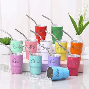 May include: A collection of colorful, personalized tumblers with stainless steel straws. Each tumbler is a different color, including red, green, yellow, and pink, and features a name in a cursive font. A blue tumbler is lying on its side.