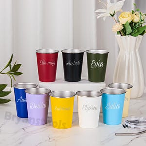 Puede incluir: Un conjunto de vasos personalizados de acero inoxidable en varios colores, como rojo, negro y azul. Cada vaso tiene un borde plateado y un nombre grabado. Un jarrón blanco con flores se encuentra en el fondo.