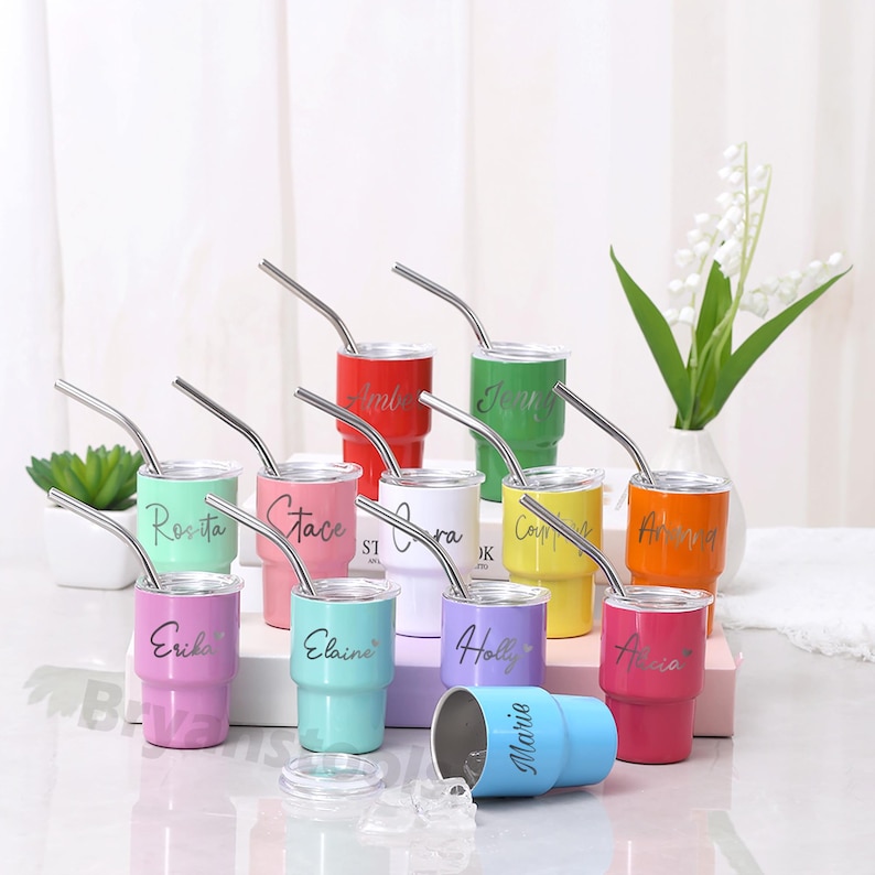 May include: A collection of colorful, personalized tumblers with stainless steel straws. Each tumbler is a different color, including red, green, yellow, and pink, and features a name in a cursive font. A blue tumbler is lying on its side.