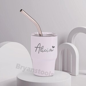 May include: A white tumbler with a silver straw. The tumbler has the name "Alicia" in script with a heart symbol. The tumbler is on a white pedestal with abstract white shapes in the background.