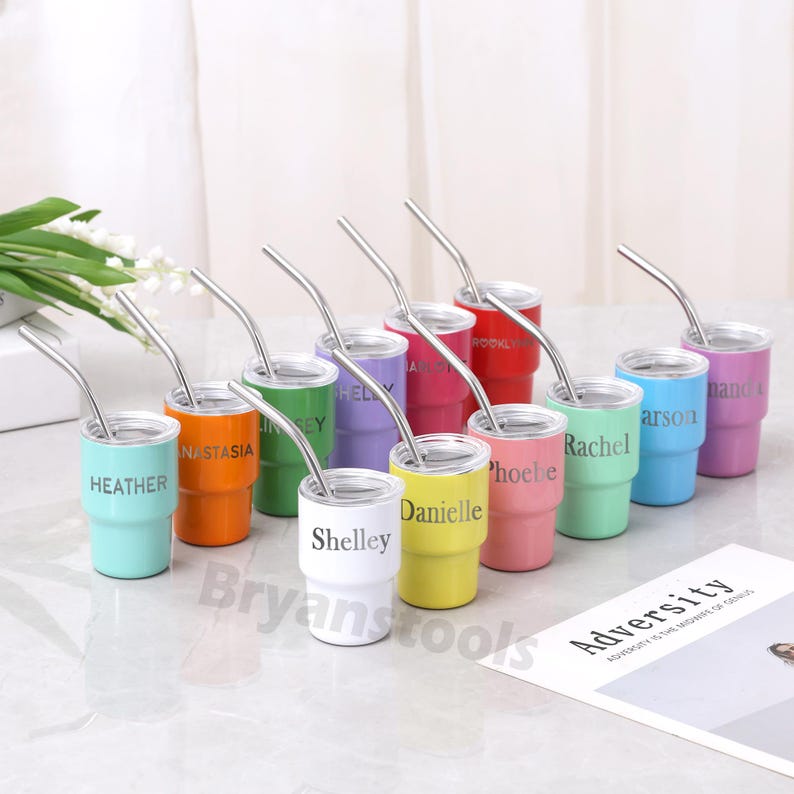 May include: A collection of colorful, personalized stainless steel tumblers with silver straws. Each tumbler is a different color and has a name printed on it. The tumblers are arranged on a white surface.