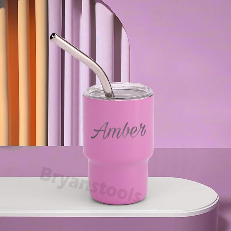May include: A pink tumbler with a silver straw and a clear lid. The name "Amber" is written on the side in a cursive font. The tumbler is on a white surface against a purple background.