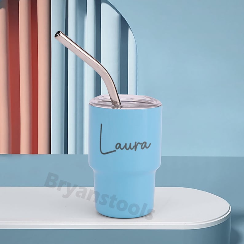 May include: A light blue tumbler with a silver straw and a clear lid. The name "Laura" is written in a cursive font on the side of the tumbler. The tumbler is on a white surface against a blue background.