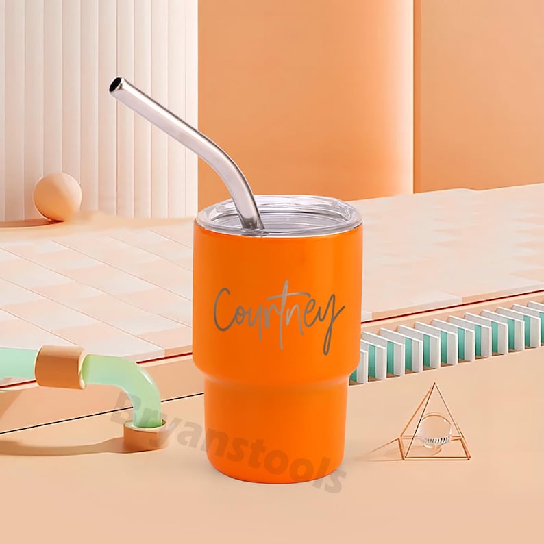 May include: An orange tumbler with a silver straw. The tumbler has the name "Courtney" in a cursive font. The tumbler is cylindrical with a slightly wider base and a silver lid. The background is a light orange.
