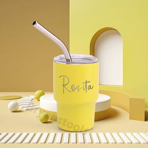 May include: A yellow tumbler with a silver straw and a clear lid. The name "Rosita" is written on the side in a cursive font. The tumbler is on a white and yellow background.