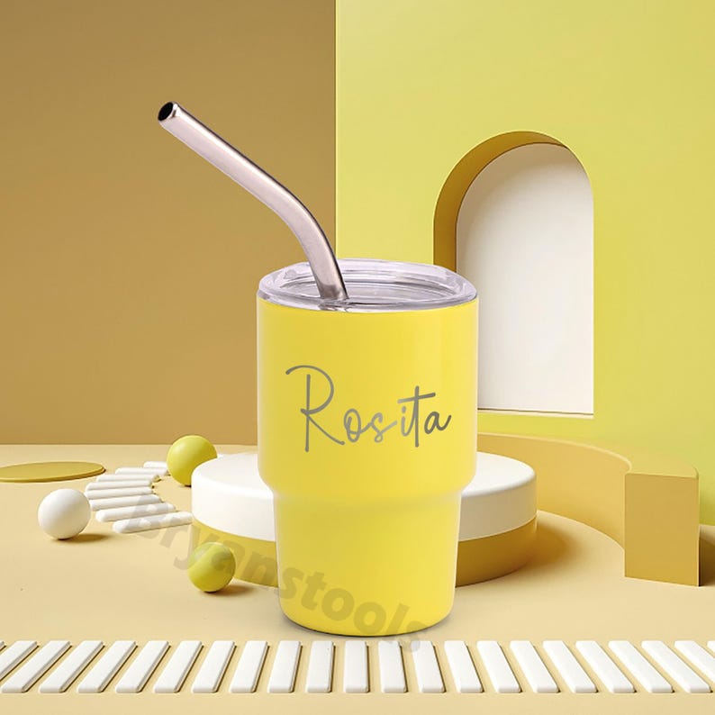 May include: A yellow tumbler with a silver straw and a clear lid. The name "Rosita" is written on the side in a cursive font. The tumbler is on a white and yellow background.