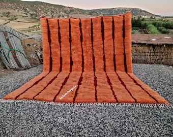 May include: An orange rug with vertical dark red stripes. The rug has a textured surface and a fringe along the bottom edge. The rug is displayed outdoors on a bed of small gray stones.