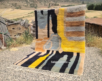Könnte beinhalten: Rechteckiger Teppich mit abstraktem Design in Gelb-, Schwarz-, Blau- und Beigetönen. Der Teppich hat einen Fransenbesatz am unteren Rand. Er wird im Freien mit einer natürlichen Landschaft im Hintergrund präsentiert.
