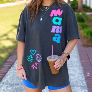 May include: Dark grey t-shirt with the word "mama" in pink, teal, and purple. The shirt also features a heart, flower, smiley face, and four small squares in teal, pink, and purple. The person is wearing blue shorts and holding a drink.