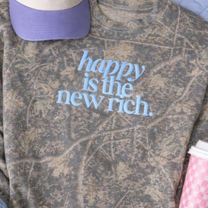 May include: A grey camouflage-patterned sweatshirt with the light blue text "happy is the new rich." A purple baseball cap is partially visible at the top left. A pink and white to-go coffee cup is visible on the right.