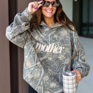 May include: A camouflage-patterned hoodie with the word "mother" in a cursive font across the chest. The hoodie is a mix of green, brown, and tan colors. The person is holding a mug with a plaid pattern.