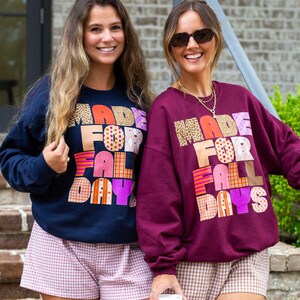 May include: A navy blue sweatshirt and a maroon sweatshirt, both featuring the text MADE FOR FALL DAYS in block letters with various patterns including leopard print, plaid, and polka dots, paired with light-colored gingham patterned shorts.