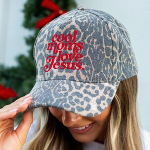 May include: A baseball cap featuring a grey and black leopard print pattern. The front of the cap displays the text "cool moms love Jesus." embroidered in red thread. The cap has a structured crown and a curved brim with matching animal print.