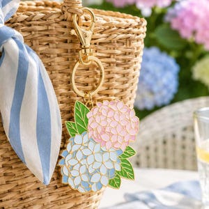 May include: A gold-toned metal keychain featuring an enamel charm of two hydrangea flower clusters, one in light blue and one in soft pink, accented with green leaves. The charm is attached to a gold-colored lobster clasp and a circular ring.