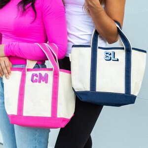 May include: Two canvas tote bags with dual handles. One bag features bright pink trim and the embroidered letters CM. The other bag has navy blue trim and the embroidered letters SL. Both bags are made of off-white canvas material.