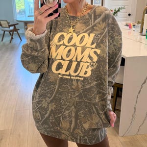 Könnte beinhalten: Ein Sweatshirt-Kleid mit Camouflage-Muster und dem Text "COOL MOMS CLUB" in gelber Schrift. Das Design beinhaltet ein Smiley-Gesicht und den Satz "ALL MOMS WELCOME". Das Kleid hat eine lockere Passform und lange Ärmel.