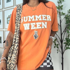 May include: An orange short-sleeved t-shirt featuring the text SUMMER WEEN in white block letters with gold glitter trim. Below the text is a patch of a skeleton hand making a peace sign. A leopard print bag strap hangs over the shoulder.