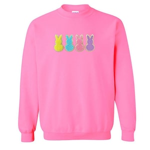 May include: A bright pink sweatshirt featuring four colorful bunny designs. The bunnies are yellow, turquoise, pink, and purple, each with a textured appearance. The sweatshirt has a crew neck and long sleeves.