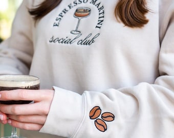 Puede incluir: Sudadera beige con el texto bordado "ESPRESSO MARTINI social club" y un gráfico de cóctel. La manga presenta tres granos de café bordados. Se sostiene una copa de cóctel en primer plano.