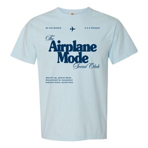 May include: Light blue t-shirt with the text "The Airplane Mode Social Club" in navy blue. The shirt also features a small airplane graphic and the phrases "do not disturb" and "it's a lifestyle."