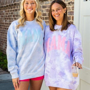 May include: Two women wearing tie-dye sweatshirts. One is light blue with "MAMA" in light blue lettering. The other is purple with "MAMA" in pink lettering. Both have crew necks and long sleeves. The women are smiling.