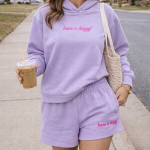 以下が含まれることがあります： ライラック色のパーカーとショートパンツのセット。パーカーとショートパンツには、ピンク色のスクリプトで「have a dayzy！」というフレーズが書かれています。ショートパンツはウエストゴム仕様です。透明なプラスチックカップに入ったコーヒーを手に持っています。