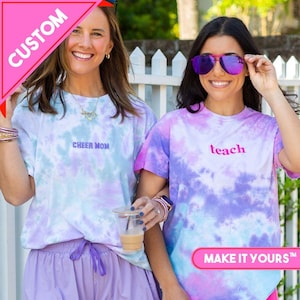 Op de afbeelding: Twee vrouwen dragen tie-dye t-shirts met de woorden "CHEER MOM" en "teach" erop geborduurd. De shirts zijn in tinten paars, blauw en wit. De afbeelding bevat ook de tekst "CUSTOM" en "MAKE IT YOURS".