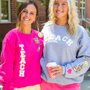 May include: A bright pink sweatshirt with the word TEACH in white letters and patches of a coffee cup, heart, pencil, and smiley face, alongside a gray sweatshirt with the word TEACH in white letters and similar patches on the sleeve.