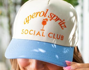 May include: A cream-colored baseball cap with a light blue brim and top button. The cap features the embroidered text "aperol spritz SOCIAL CLUB" in orange, along with a cocktail glass graphic.