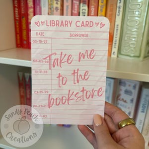 May include: A pink and white library card with the text "Take me to the bookstore" in pink script. The card has a heart design and the words "Library Card" at the top. The card is held in front of a bookshelf.