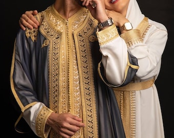Può includere: Una coppia in abiti tradizionali, l'uomo indossa una tunica nera e dorata ricamata e la donna indossa un abito bianco e dorato ricamato e un velo bianco. Sorridono e si guardano.