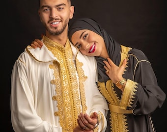 Può includere: Una coppia vestita con abiti tradizionali marocchini. L'uomo indossa una djellaba bianca con ricami dorati e la donna indossa una djellaba nera con ricami dorati e un velo nero.