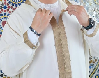 Può includere: Un capo color crema a maniche lunghe con finiture e dettagli dorati. Il capo ha una fascia dorata decorativa sul davanti e sui polsini. Lo sfondo presenta un colorato motivo a mosaico.