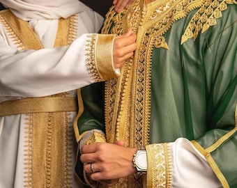 Può includere: Una coppia sorridente, vestita con abiti tradizionali. La donna indossa un abito bianco e un velo con finiture dorate. L'uomo indossa una veste verde con ricami dorati e maniche bianche. Lo sfondo è nero.
