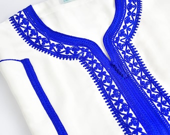 Könnte beinhalten: Weißes Tunika mit aufwendiger blauer Stickerei um den Halsausschnitt und die Vorderseite. Die Tunika hat einen V-Ausschnitt und einen blauen Streifen an der Seite. Das Etikett lautet "Caftan Nawal".