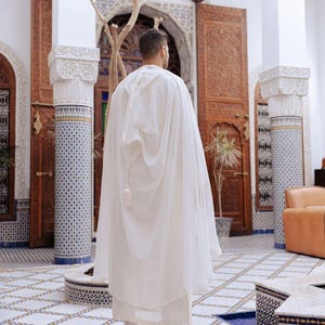 May include: A person wearing a white hooded cloak and matching pants, standing in a courtyard. The courtyard features ornate wooden doors, decorative pillars, and a patterned tile floor. The overall aesthetic suggests a cultural or historical setting.