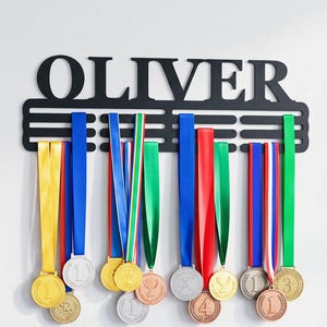 May include: A black metal wall-mounted medal display hanger featuring the name OLIVER in large capital letters at the top. The rack has three horizontal bars for hanging multiple medals with colourful ribbons in yellow, blue, red, green, and white.