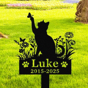 Puede incluir: Estaca de jardín de metal negro con silueta de gato que alcanza una mariposa. La estaca incluye el nombre "Luke" y los años "2015-2025". Detalles florales y huellas de patas rodean el nombre.