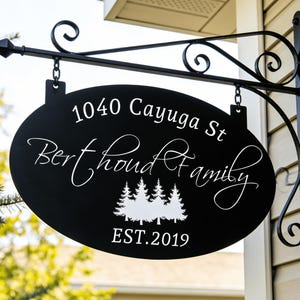 May include: Black oval sign with white text and a decorative metal bracket. The sign reads "1040 Cayuga St Berthoud Family EST.2019" with a graphic of three pine trees. The sign is hanging outside a house.