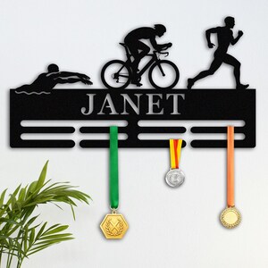 May include: Black metal medal display with the name "JANET" in silver lettering. Silhouettes of a swimmer, cyclist, and runner are above the name. Three medals with colourful ribbons hang below.