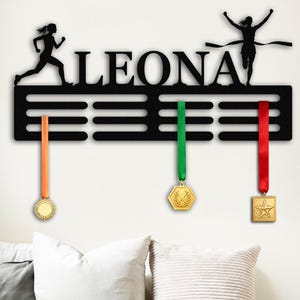以下が含まれることがあります： 「LEONA」という名前が切り抜かれた黒い金属製のメダルディスプレイ。名前の上にはランナーのシルエットがあります。オレンジ、緑、赤のリボンが付いた3つのメダルが下にぶら下がっています。メダルは金色で、さまざまな形をしています。