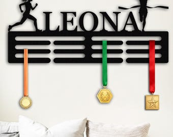 Puede incluir: Expositor de medallas de metal negro con el nombre "LEONA" recortado. Siluetas de corredores están encima del nombre. Tres medallas con cintas naranja, verde y roja cuelgan debajo. Las medallas son doradas y tienen diferentes formas.