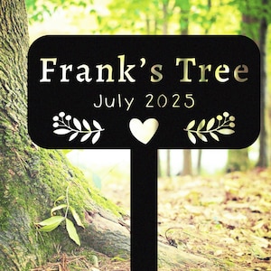 May include: Black metal tree marker with the text "Frank's Tree" in gold lettering, along with the date "July 2025". The marker features a heart and leaf designs. The marker is placed in front of a tree.