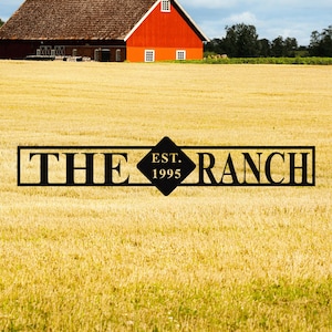 Pode incluir: Uma placa de metal preta com as palavras "THE RANCH EST. 1995" é colocada em um fundo de um campo dourado e um celeiro vermelho. O celeiro tem um telhado marrom e molduras de janelas brancas. O céu é azul.