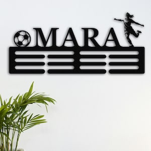以下が含まれることがあります： 黒い金属製のウォールアート。名前「MARA」、サッカーボール、サッカー選手がボールを蹴るシルエットが特徴です。名前は3本の水平バーの上にあり、装飾的なスポーツをテーマにした作品になっています。